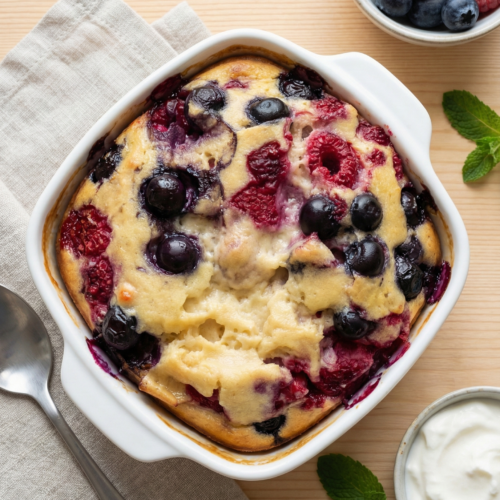 Draufsicht auf einen frisch gebackenen Beerenauflauf mit Blaubeeren und Himbeeren in einer weißen quadratischen Auflaufform, serviert auf einem Holztisch mit einer Schale Skyr, frischen Beeren, Minze und einem Löffel auf einem Leinentuch. Perfekt für ein gesundes Frühstück oder Dessert.