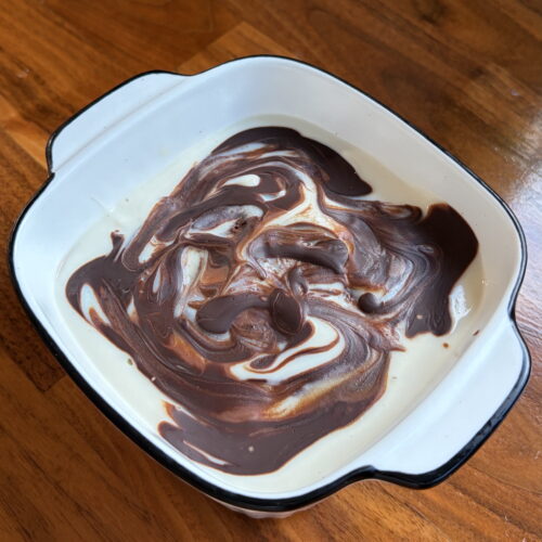 Cremiges Joghurt-Dessert mit dunklem Schokoladen-Swirl serviert in einer weiß-schwarzen Auflaufform auf Holztisch.