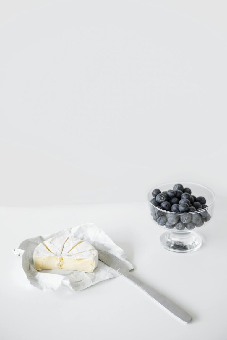 A minimalist still life featuring a glass bowl of blueberries and a cheese wedge on a white background.