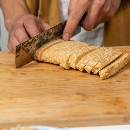 Hände schneiden einen Block Tempeh mit einem großen Messer auf einem Holzbrett in Scheiben, im Vordergrund eine unscharfe Schale mit Kichererbsen.