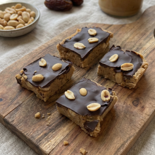 Gesunde No-Bake Erdnussbutter-Riegel mit Datteln und einer knackigen Schicht aus dunkler Schokolade, serviert auf einem rustikalen Holzbrett.