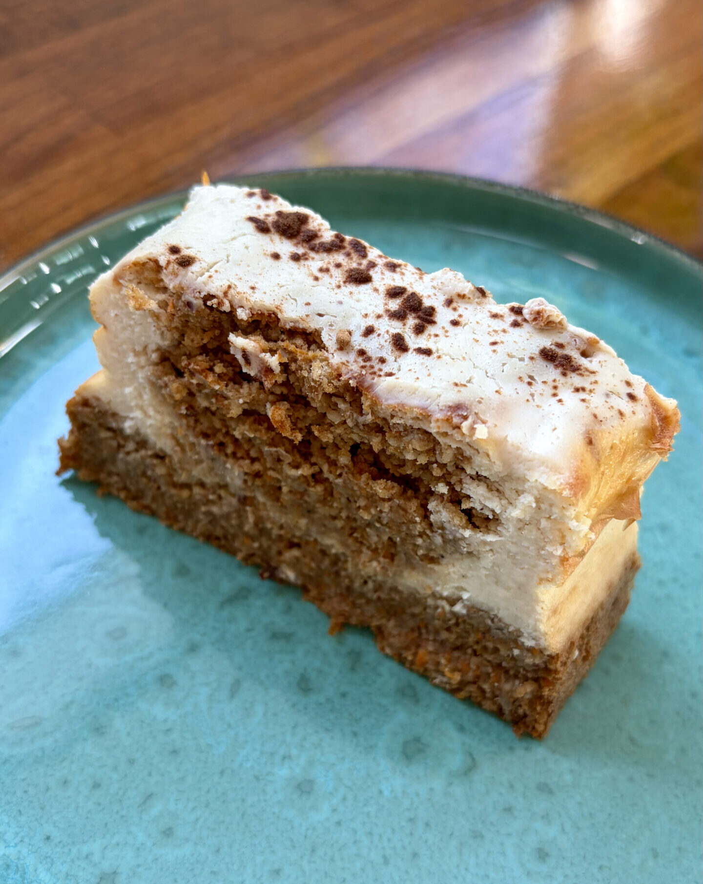 Saftiges Stück Karottenkuchen mit dicker weißer Frischkäse-Glasur und feiner Zimt-Bestäubung auf einem türkisblauen Keramikteller.