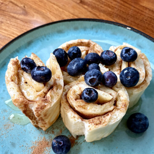 Drei aufgerollte Pfannkuchen-Röllchen auf einem türkisfarbenen Teller, bestreut mit Zimt und garniert mit frischen Blaubeeren.