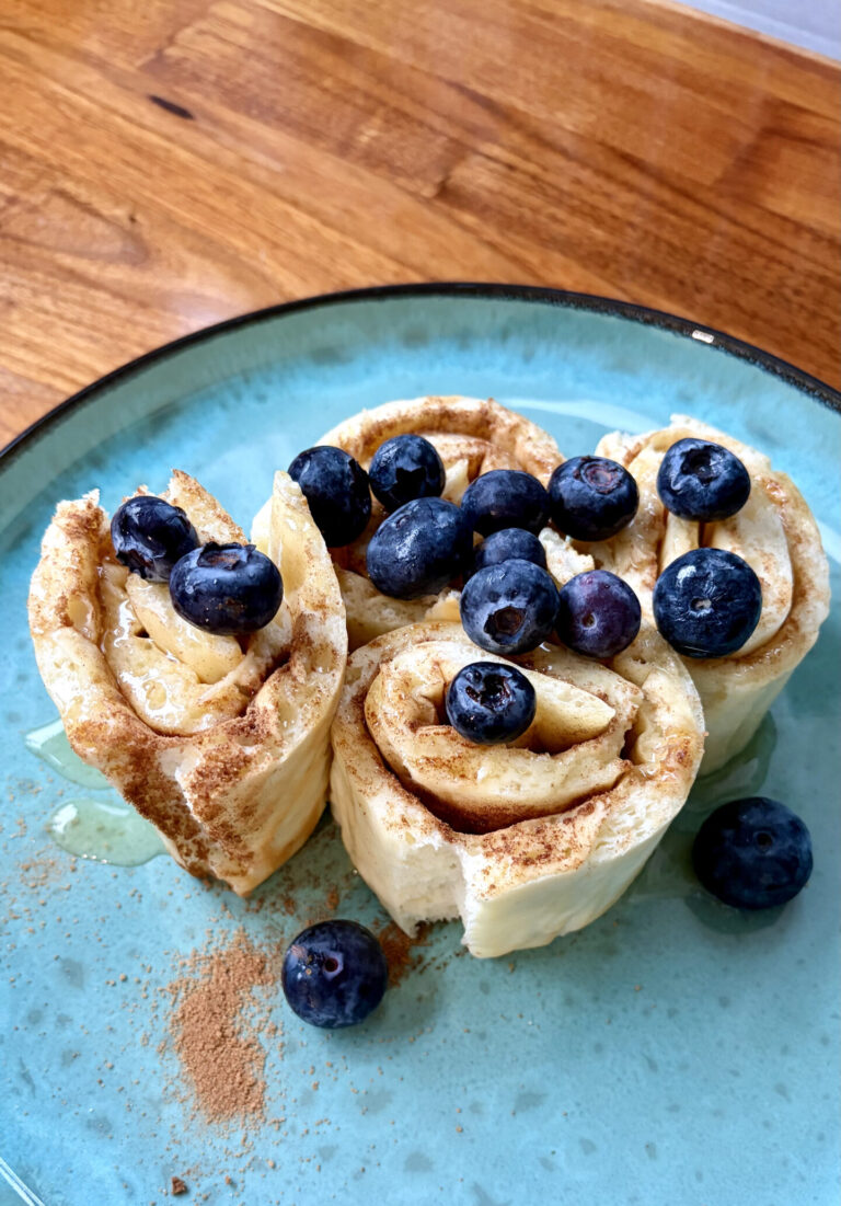Drei aufgerollte Pfannkuchen-Röllchen auf einem türkisfarbenen Teller, bestreut mit Zimt und garniert mit frischen Blaubeeren.